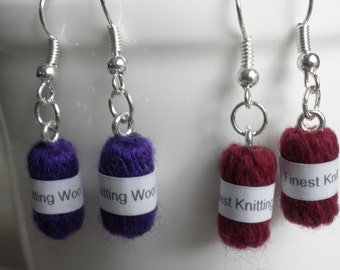 May include: Pair of dangle earrings featuring small yarn balls with labels that read "Finest Knitting Wool" and "Knitting Wool". The yarn balls are purple and burgundy.