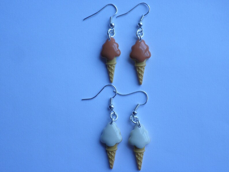 May include: A pair of silver earrings with ice cream cone charms. One pair features a brown and white ice cream cone charm, and the other pair features a white and yellow ice cream cone charm.