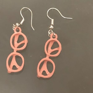 May include: A pair of pink dangle earrings shaped like glasses. The earrings have silver hooks.