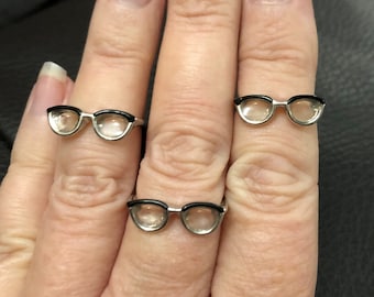 May include: Three silver rings with black rims and clear glass lenses shaped like eyeglasses.