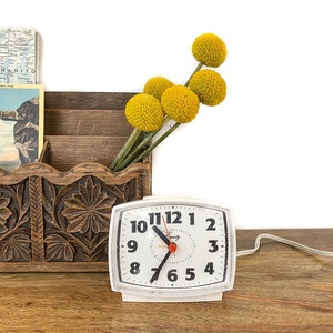 May include: White vintage alarm clock with black numbers and an orange second hand. The clock is sitting on a wooden surface with a carved wooden box and yellow flowers in the background.