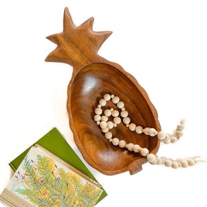 Può includere: Una ciotola di legno a forma di ananas con una finitura in legno naturale. La ciotola è piena di una collana di perline di legno bianche.