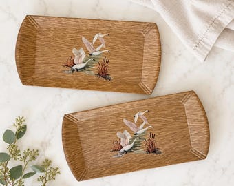 May include: Two rectangular wooden trays featuring a natural wood grain finish. Each tray displays a painted illustration of white swans swimming in water surrounded by brown reeds and cattails, with angled corners on the edges.
