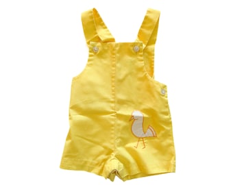 May include: Yellow baby overalls with a white bird embroidered on the front. The overalls have adjustable straps and button closures.
