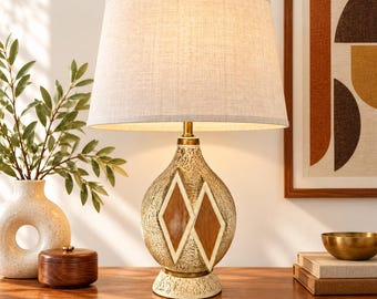 May include: A table lamp with a textured, cream-colored ceramic base featuring two brown diamond-shaped inlays and a brass-toned neck. The lamp is topped with a tapered, light-colored fabric drum shade that emits a warm, soft glow when illuminated.