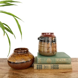May include: Two ceramic vases, one with a handle, on a stack of books. The vases are brown, black, and red with a speckled glaze. The books are green and have gold lettering. The books are titled "The Veils of  Muel Over", "Kentucky Derby Winner", and "Grosset & Dunlap".