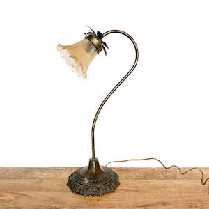 Puede incluir: Una lámpara de escritorio de estilo vintage con un tallo curvo de color latón y una base decorativa. La lámpara presenta una pantalla de vidrio ámbar en forma de campana con un borde con volantes. La lámpara está diseñada para un escritorio o mesa y tiene un cable de alimentación.