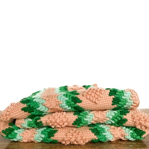 May include: A folded crocheted blanket with a pink and green geometric pattern. The blanket is made of soft, fluffy yarn and has a textured surface.