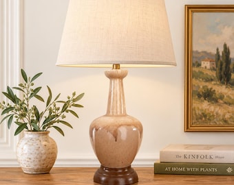 May include: A table lamp with a beige ceramic base featuring a glossy drip glaze finish, a dark brown circular wooden pedestal, and a tapered off-white fabric lampshade topped with a small gold-toned finial, standing on a wooden surface.