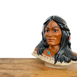 Könnte beinhalten: Eine Keramikbüste einer indianischen Frau mit langen schwarzen Haaren, die eine türkisfarbene Halskette und rote Ohrringe trägt. Die Büste ist in Braun-, Beige- und Schwarztönen bemalt.