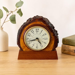 Puede incluir: Un reloj de sobremesa con esfera circular, números arábigos y manecillas negras, enmarcado en madera natural con bordes de corteza oscura y rugosa. El reloj está montado sobre una base rectangular de madera lisa de color marrón medio.