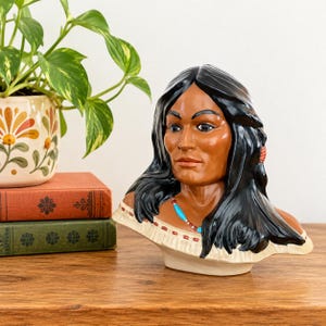 May include: A ceramic bust of a person with long black hair, tan skin, and dark eyes. The figure wears an off-white garment with a fringed neckline and a beaded necklace featuring small blue and red beads. The sculpture has a smooth finish.