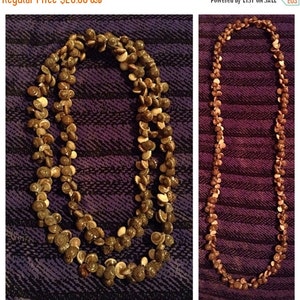 May include: Two necklaces made of small, natural seashells. The shells are primarily brown and beige, strung together to form a long, layered necklace. The background is a dark purple fabric.