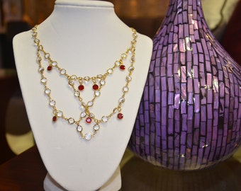 May include: A gold necklace with a layered design features clear and red round stones. The necklace is displayed on a white mannequin bust.