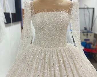 May include: A white wedding dress with a sheer bodice and long sleeves. The dress is covered in sequins and beads.