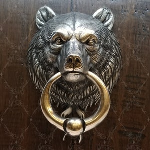 Può includere: Un batacchio per porta a forma di testa d'orso argentata con un anello dorato. L'orso ha una texture di pelliccia realistica e un viso dettagliato.