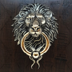 May include: A gold lion's head door knocker with a ring handle. The knocker is mounted on a dark wooden door.