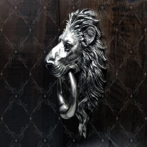 May include: A silver lion's head door knocker with a ring. The knocker is mounted on a wooden door.