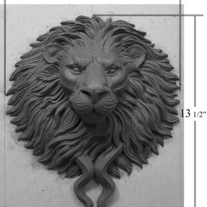 Pode incluir: Escultura de cabe&ccedil;a de le&atilde;o em tons de cinza, medindo 29 cm de largura e 34 cm de altura. A escultura apresenta uma juba detalhada, um nariz proeminente e elementos curvos estilizados na base. A express&atilde;o do le&atilde;o &eacute; estoica.