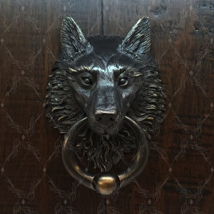 Puede incluir: Un llamador de puerta con forma de cabeza de lobo con acabado en bronce. La boca del lobo está abierta y el llamador tiene un anillo para golpear.