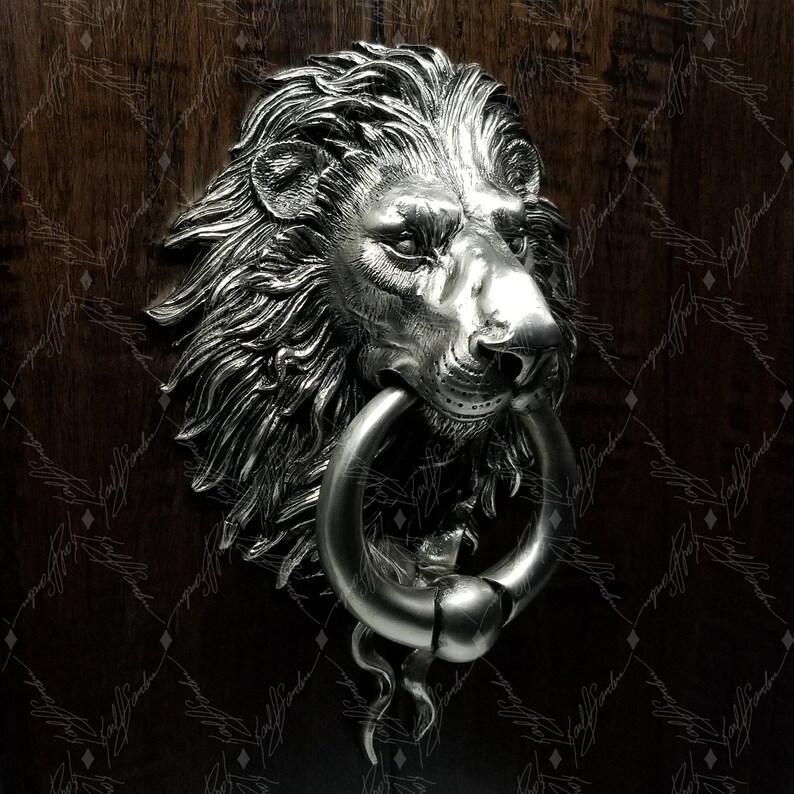 May include: A silver lion's head door knocker with a ring handle. The lion's head is detailed with a mane and a realistic expression.