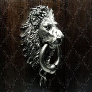 May include: A silver lion head door knocker with a ring handle. The knocker is mounted on a wooden door.