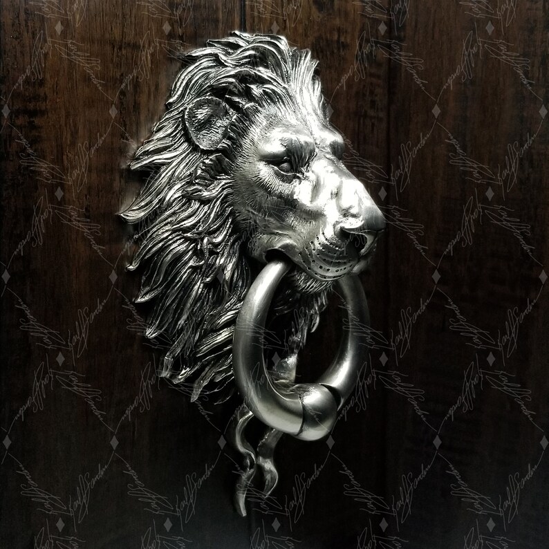 May include: A silver lion head door knocker with a ring handle. The knocker is mounted on a wooden door.