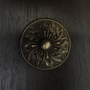 May include: A circular, ornate, brass door knob with a floral design. The knob has a raised center and is set against a dark wood background.