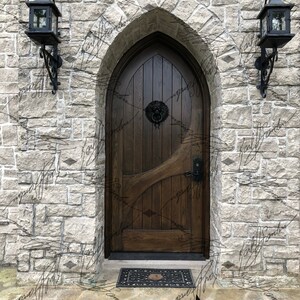 Pode incluir: Uma porta arqueada de madeira escura com uma aldrava em forma de cabe&ccedil;a de le&atilde;o est&aacute; situada numa parede de pedra. Duas lanternas pretas penduram-se em ambos os lados da porta. Um gato preto est&aacute; sentado no p&aacute;tio de pedra em frente &agrave; porta.