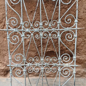 Op de afbeelding: Een decoratief metalen hek met een traditioneel Marokkaans ontwerp. Het hek is gemaakt van smeedijzer en heeft ingewikkelde krulwerk en een lichtgrijze afwerking.