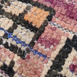 May include: Close-up of a woven rug with a geometric pattern in shades of pink, orange, white, and black. The rug has a textured, hand-woven look.