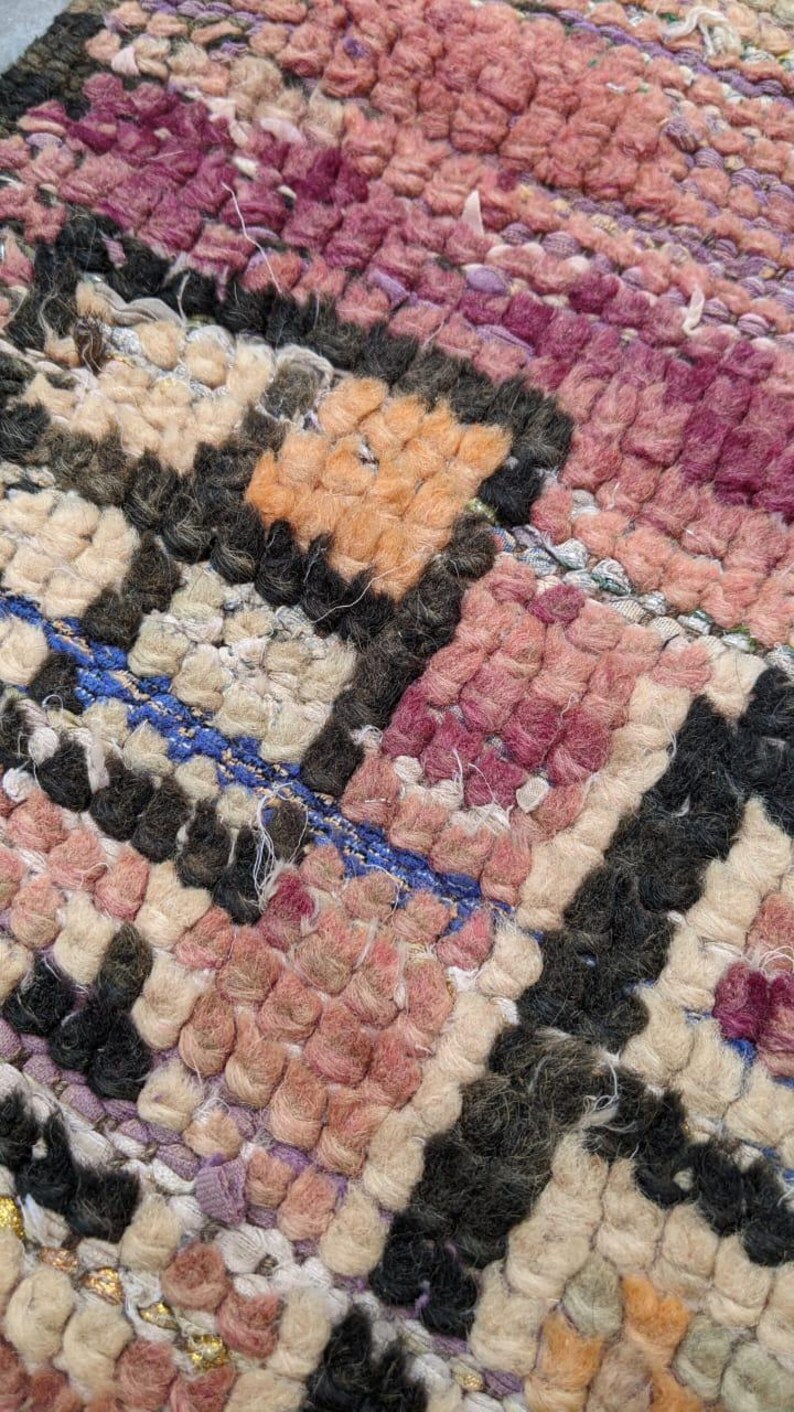 May include: Close-up of a woven rug with a geometric pattern in shades of pink, orange, white, and black. The rug has a textured, hand-woven look.