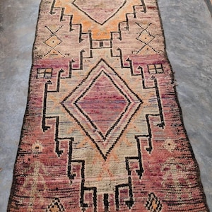 May include: A vintage Moroccan rug with a geometric pattern in shades of pink, beige, and black. The rug has a fringed edge and is made of wool.