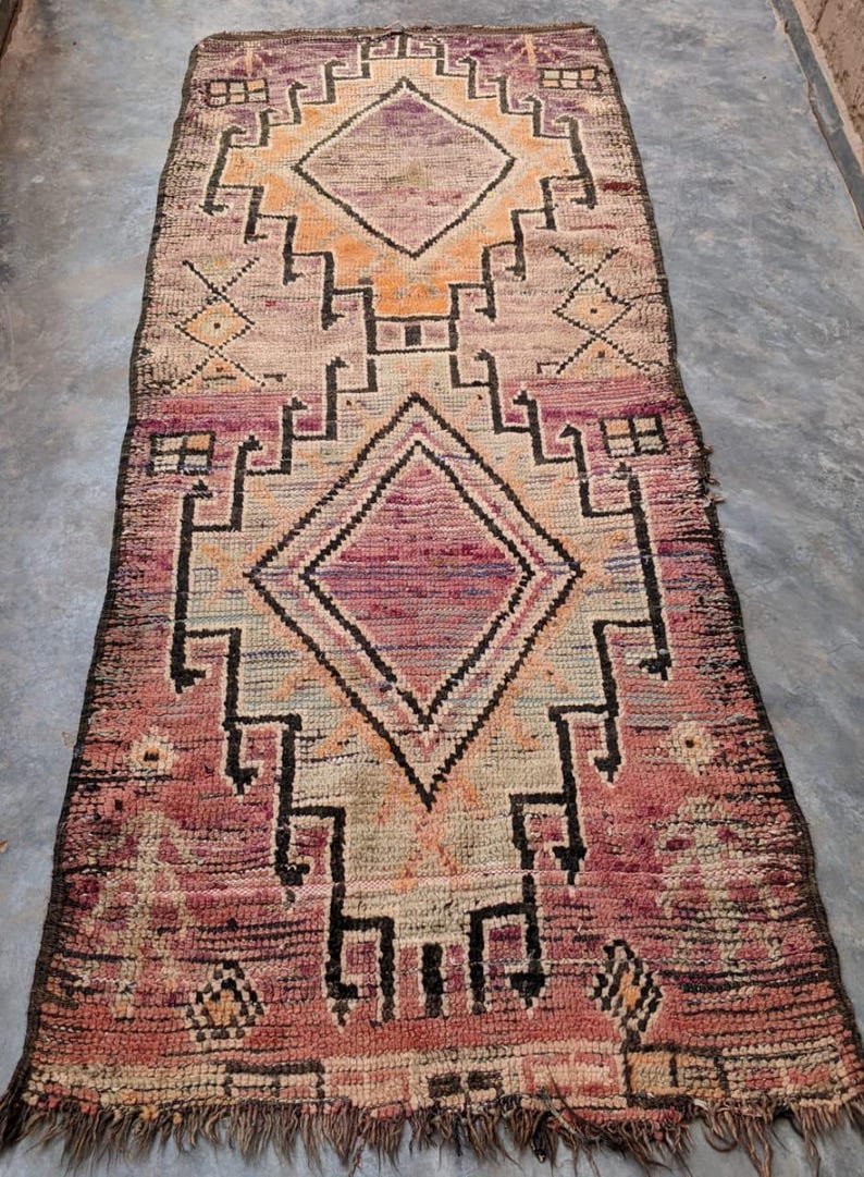 May include: A vintage Moroccan rug with a geometric pattern in shades of pink, beige, and black. The rug has a fringed edge and is made of wool.