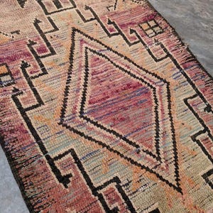May include: A vintage Moroccan rug with a geometric pattern in shades of pink, orange, and black. The rug has a distressed look and is woven with a variety of textures.
