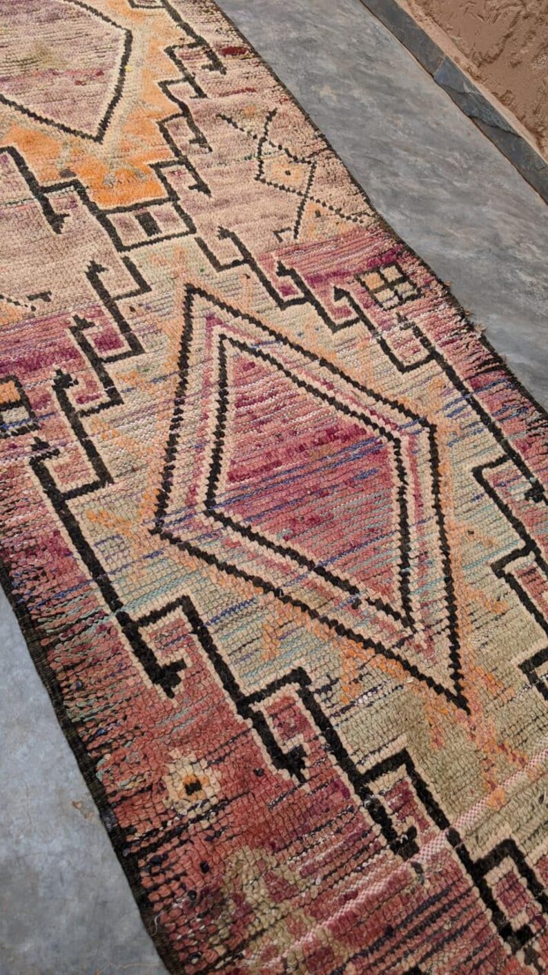 May include: A vintage Moroccan rug with a geometric pattern in shades of pink, orange, and black. The rug has a distressed look and is woven with a variety of textures.