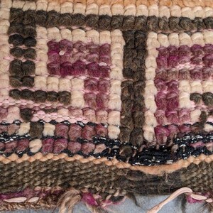 May include: Close-up of a woven rug with a geometric pattern in shades of brown, pink, and beige. The rug has a frayed edge and is made of wool.