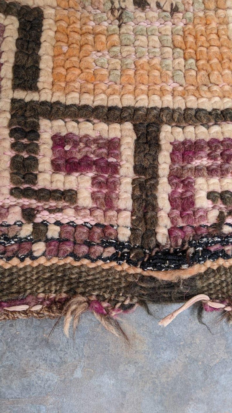 May include: Close-up of a woven rug with a geometric pattern in shades of brown, pink, and beige. The rug has a frayed edge and is made of wool.
