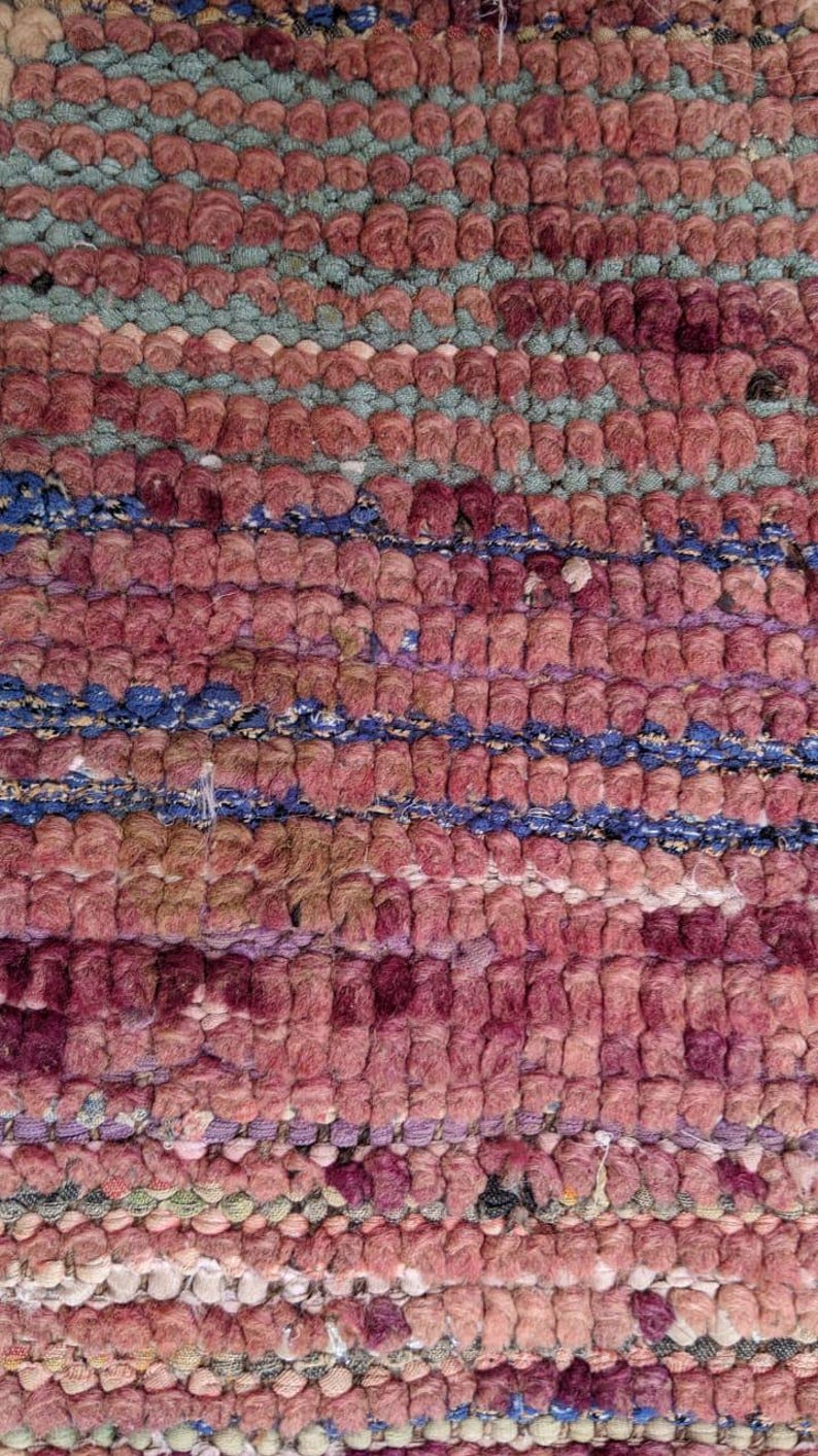 May include: Close-up of a woven rug with a pink, blue, and white pattern. The rug is made of thick, textured yarn and has a slightly distressed look.