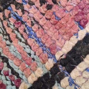 May include: Close-up of a colorful woven rug with a variety of colors including pink, blue, black, and white. The rug is made of thick, fuzzy yarn and has a textured surface.