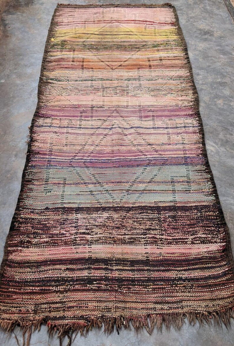 May include: A long, narrow, woven rug with a colorful striped pattern. The rug has a fringe on both ends and is made of natural fibers. The colors in the rug are a mix of pink, purple, brown, and yellow.