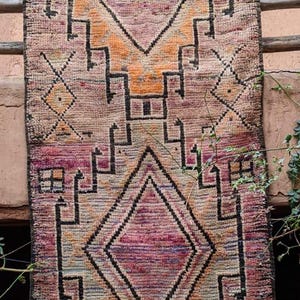 May include: A vintage Moroccan rug with a geometric pattern in shades of pink, orange, and black. The rug has a fringe border and is woven with a traditional design.