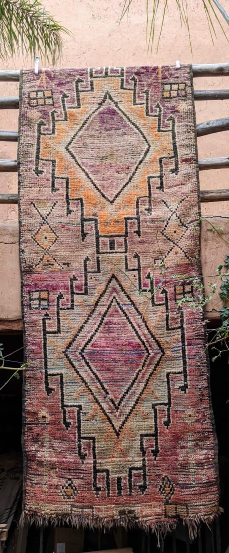 May include: A vintage Moroccan rug with a geometric pattern in shades of pink, orange, and black. The rug has a fringe border and is woven with a traditional design.