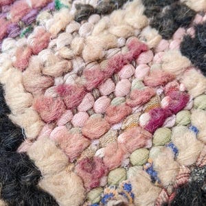 May include: Close-up of a colorful woven rug with pink, beige, and black yarn. The rug has a textured, hand-woven look.