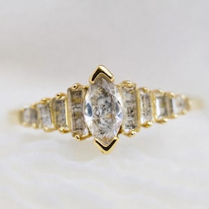 May include: A gold ring featuring a central marquise-cut salt and pepper diamond flanked by a series of smaller baguette-cut salt and pepper diamonds on each side, all set in a yellow gold band with a polished finish and prong settings.