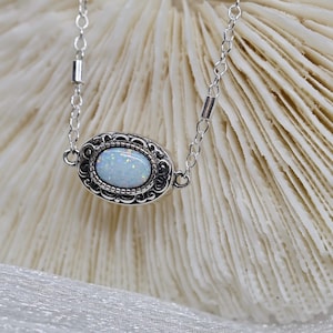 May include: A silver chain necklace with a delicate oval pendant featuring a white opal stone set in an ornate silver frame.