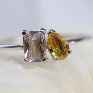 Può includere: Un anello d'argento con due pietre preziose: una pietra rettangolare marrone chiaro e una pietra gialla a forma di goccia. L'anello ha una sottile fascia d'argento e le pietre sono vicine.