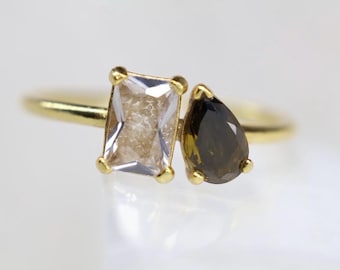 Puede incluir: Un anillo de oro con dos piedras preciosas distintas colocadas una al lado de la otra. Una piedra es transparente y rectangular con corte esmeralda, mientras que la otra es de color marrón oscuro y forma de pera. Ambas sujetas por garras de oro.