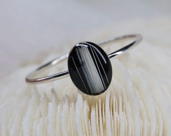 May include: A thin silver ring featuring a polished oval-shaped gemstone. The stone is primarily black with distinct, vertical white stripes running through the center, creating a high-contrast pattern set on a simple, smooth, metallic band.