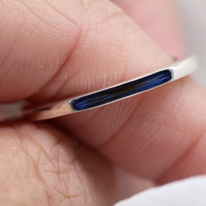 May include: A thin, polished silver-toned metal ring featuring a narrow, elongated inlay of deep blue enamel. The band has a smooth, rounded profile and a reflective surface, showcasing a minimalist design with a vibrant, dark blue color accent.